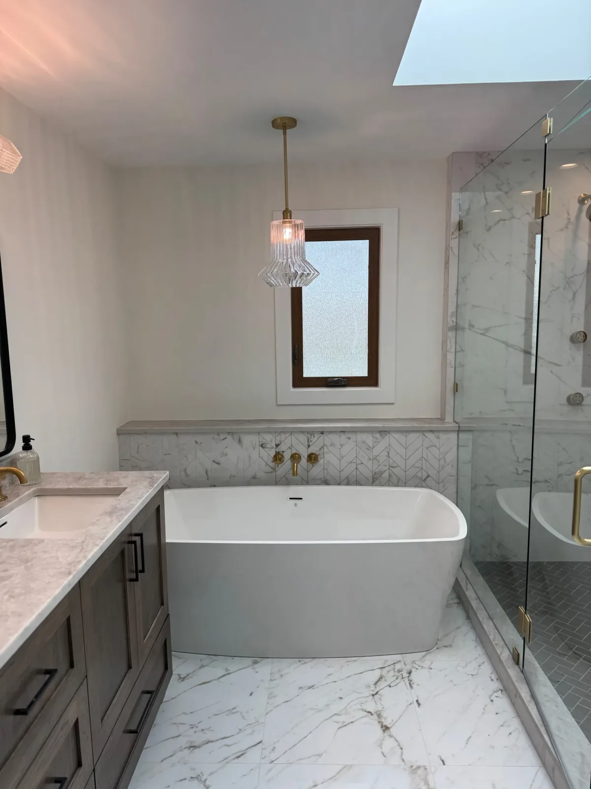 Freestanding tub in a marble-clad alcove under a fluted glass pendant, with wall-mounted brass filler and a frameless glass shower alongside - Mokena IL