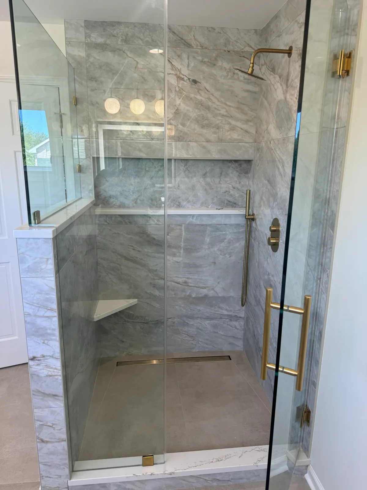 Walk-in shower lined in book-matched grey and white marble with a frameless glass door, brass rain head, corner bench and linear drain - Shorewood IL