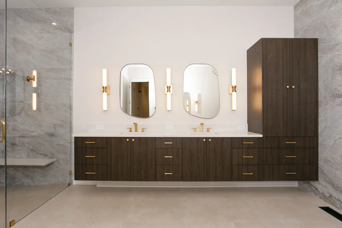 Dark walnut double vanity with a stone top, two undermount basins, brass wall-mounted faucets, arched mirrors and vertical brass sconces - Lemont IL