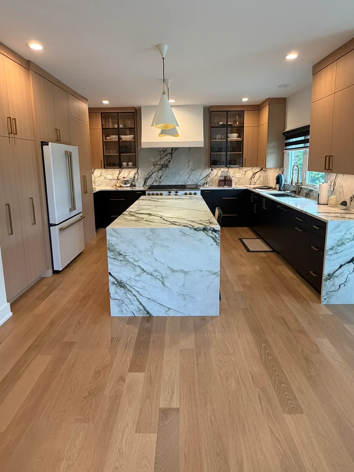Kitchen with a waterfall island in veined white and green marble, white oak cabinetry with brass pulls, glass-front uppers and a full-height marble slab behind the range - Mokena IL