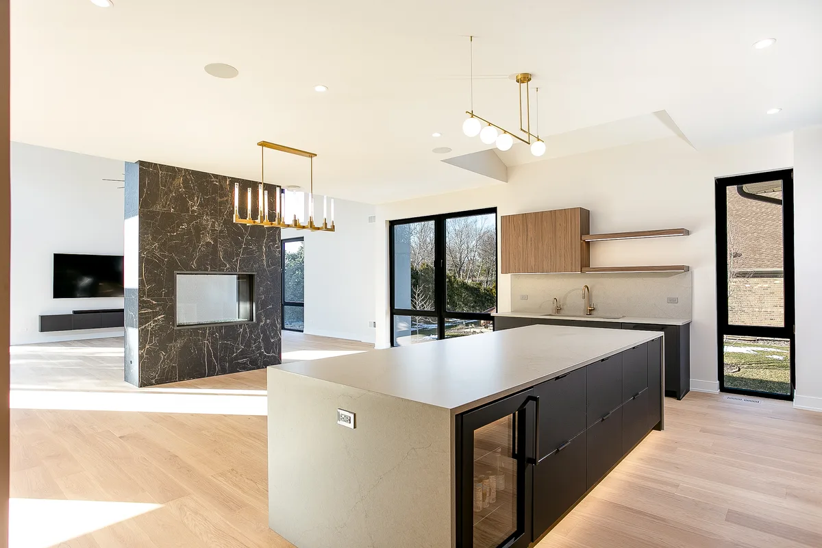 Kitchen and great room with a quartz waterfall island, a dark marble fireplace column beyond and black-framed windows - Lemont IL