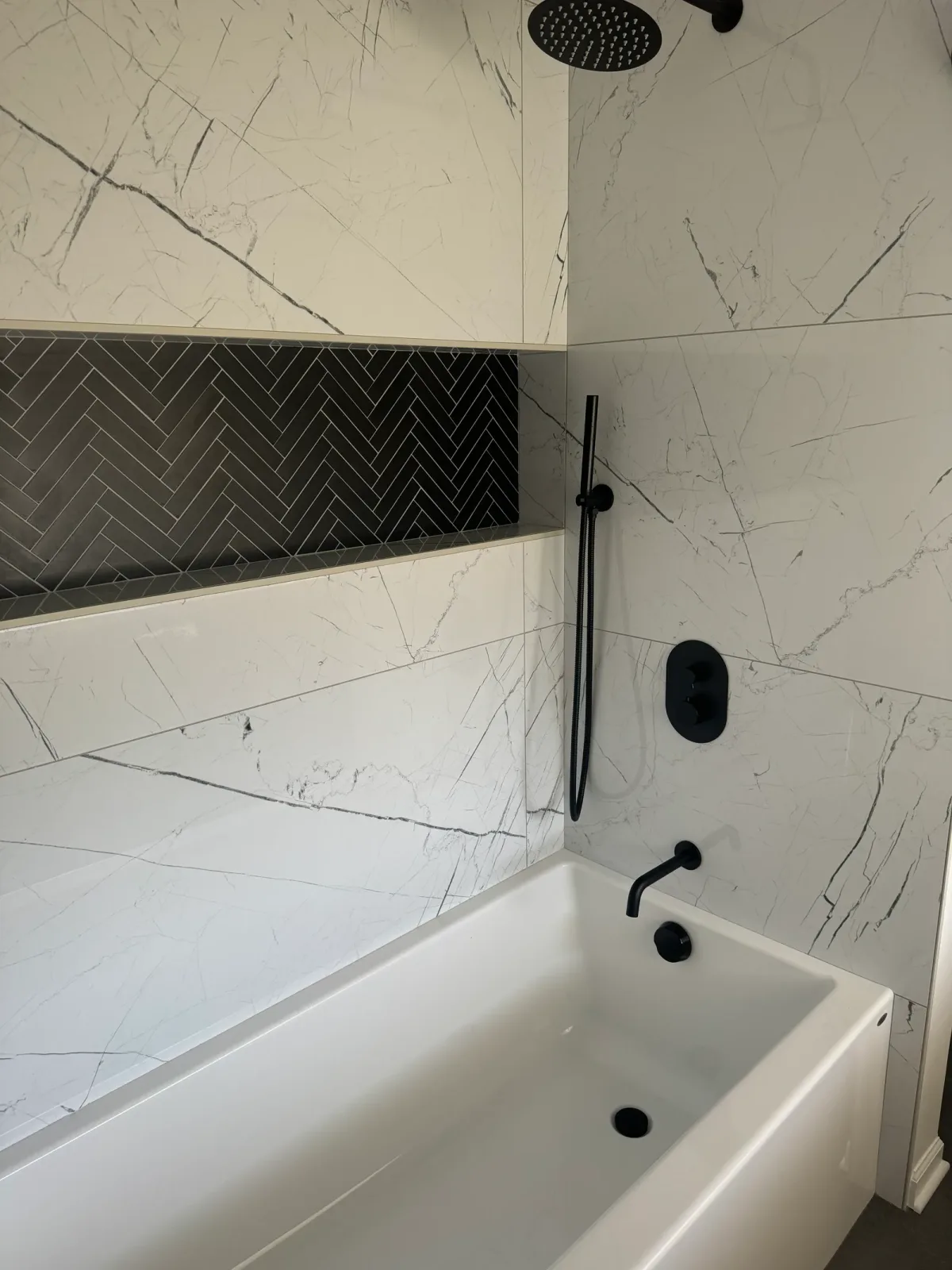 Guest bathroom remodel after photo, Shorewood IL - alcove tub in large-format white marble-look porcelain with a black herringbone niche, a matte-black handshower, control plate and wall spout