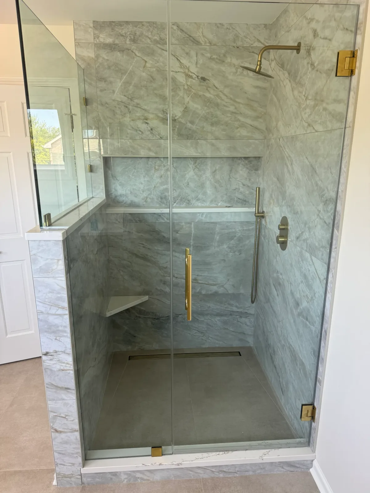 Shower remodel after photo, Shorewood IL - frameless glass shower lined in large-format grey marble-look porcelain with a brass rain head, a brass handshower and a brass ladder pull