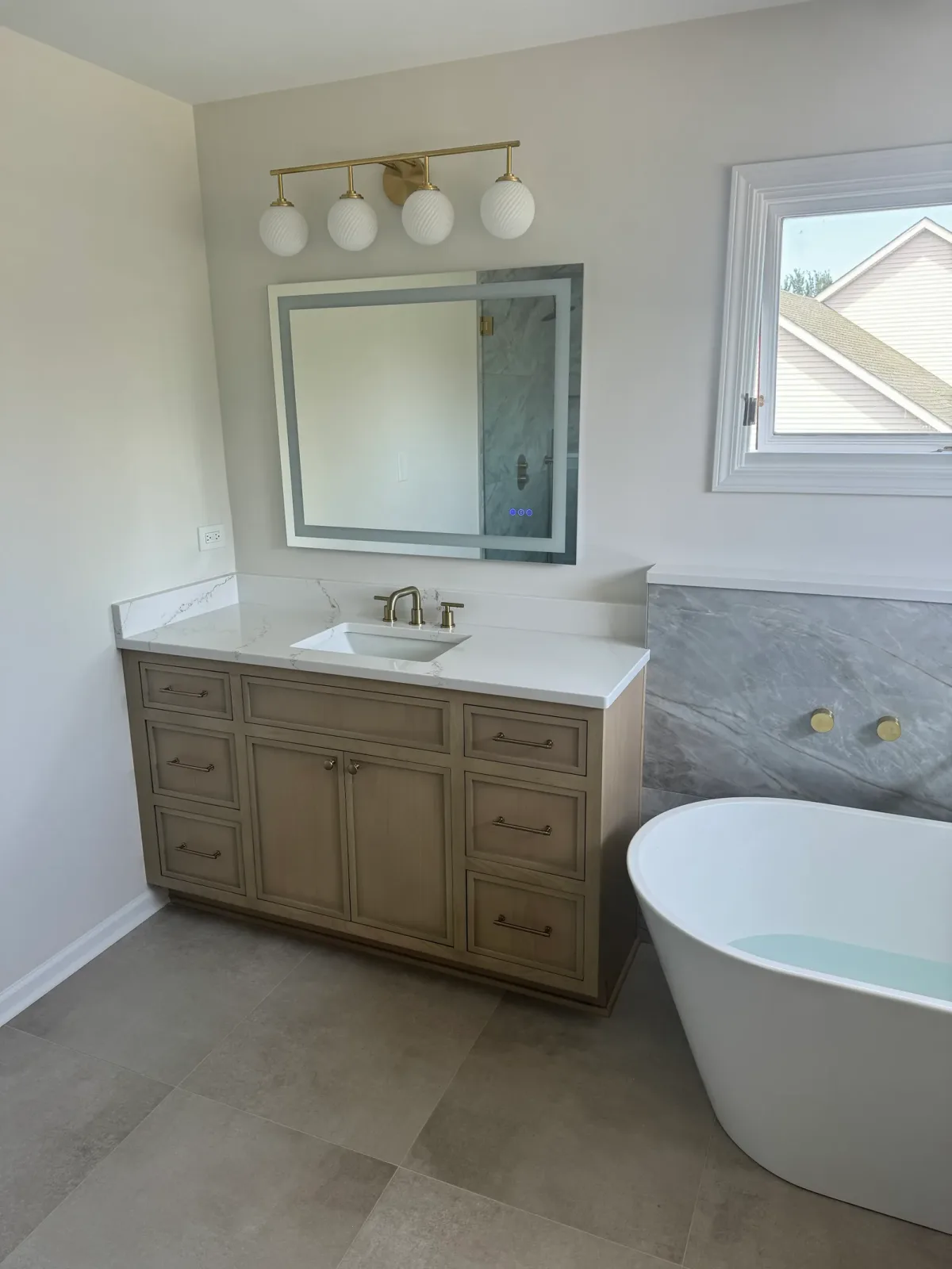 Primary bathroom remodel after photo, Shorewood IL - light-oak vanity with a white quartz top, a rectangular undermount basin, brass tapware and a backlit mirror, with the freestanding tub at right
