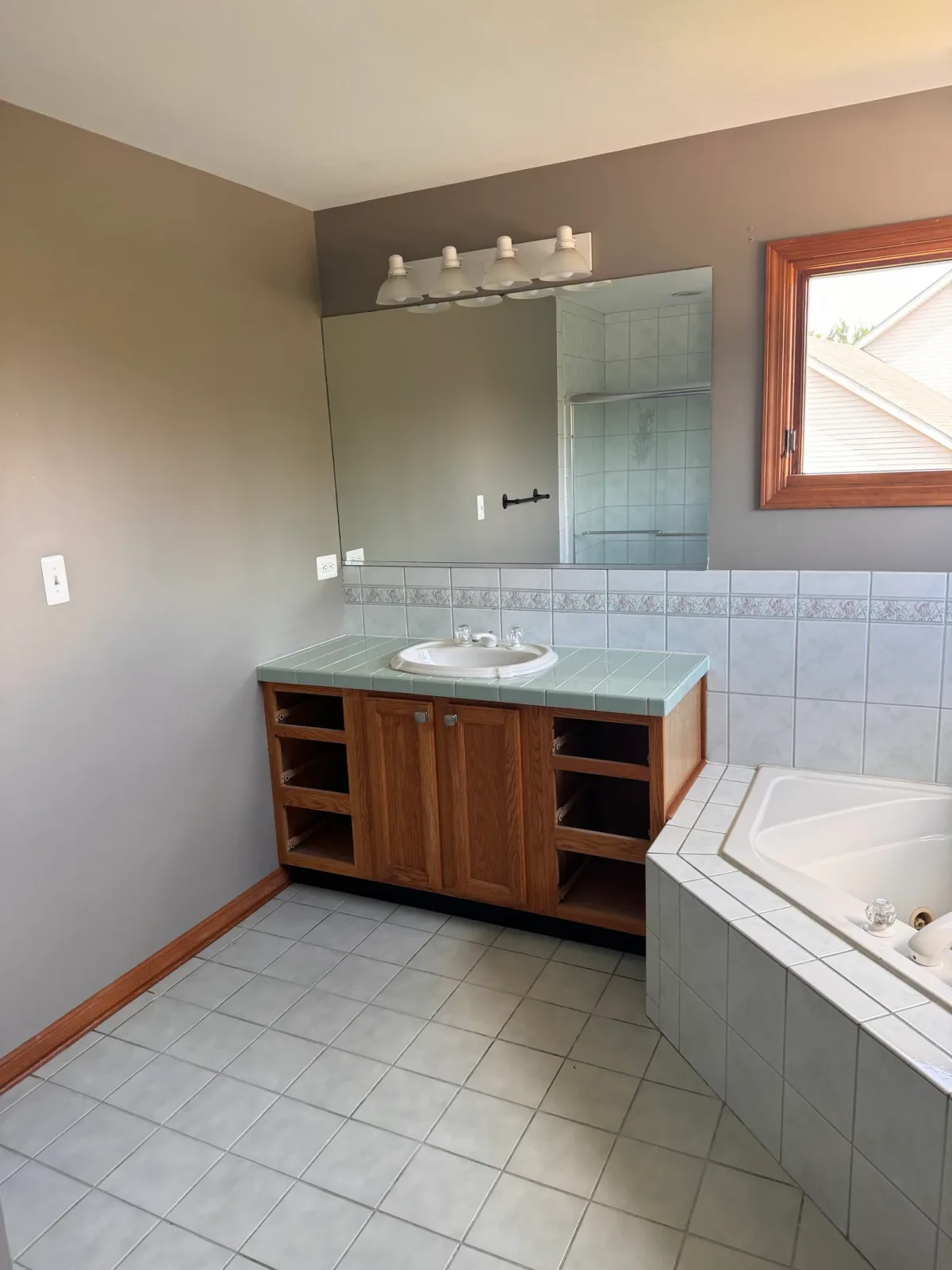 Primary bathroom before photo, Shorewood IL - oak vanity with a seafoam-green tile countertop, an oval drop-in sink and a frameless plate mirror, with the corner jetted tub at right