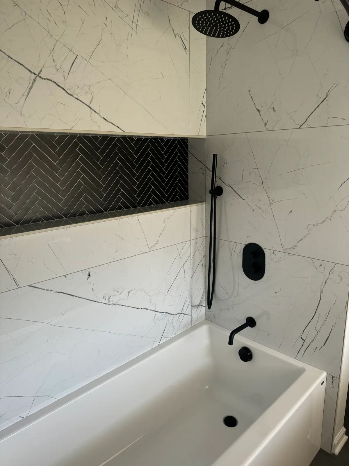 Matte-black handshower on a slide bar and a matte-black control plate set against white marble-look porcelain, below a long black herringbone mosaic niche - Shorewood IL