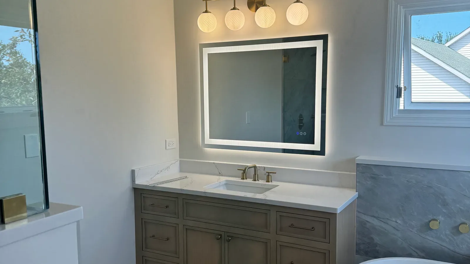 Primary bathroom in a bathroom and laundry renovation in Shorewood, IL, with a light-oak vanity, a white quartz top with grey veining, a backlit mirror, brass globe lighting and a grey marble-look porcelain tub wall by Luxe Development