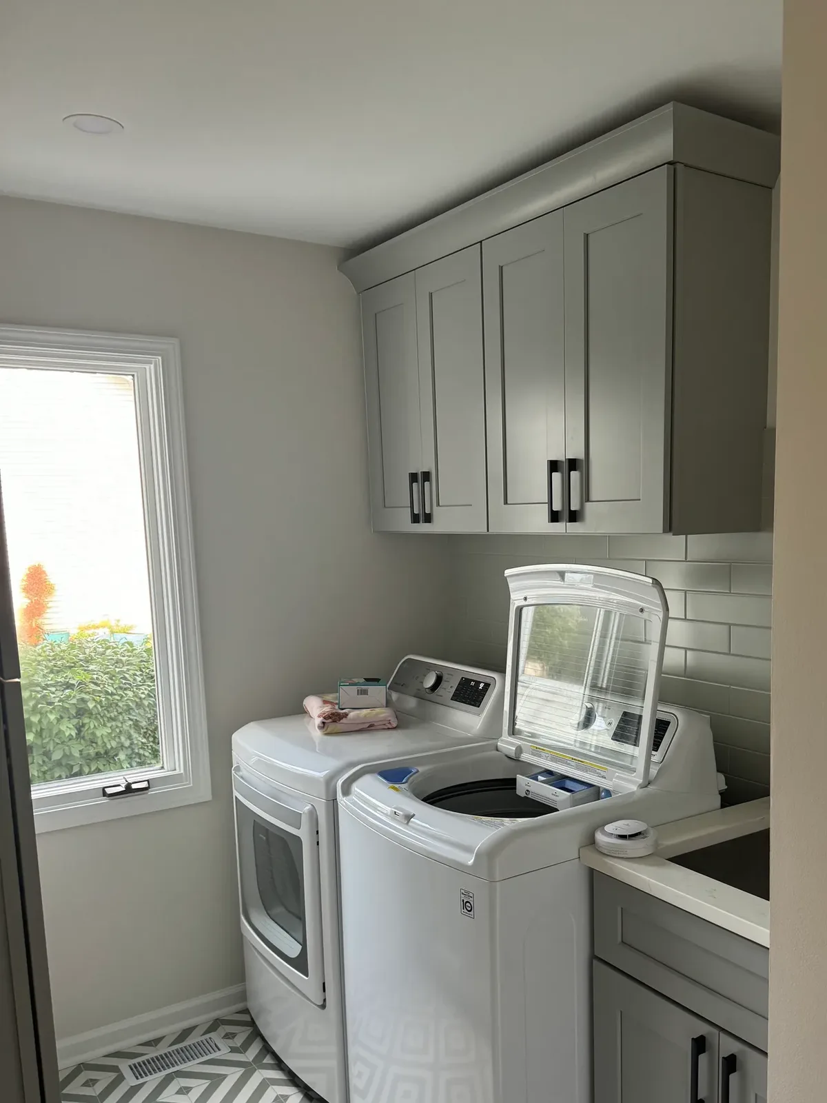 Laundry room with sage-grey shaker cabinetry over a washer and dryer, a wall cabinet run with matte-black pulls, a window and a patterned grey and white floor - Shorewood IL