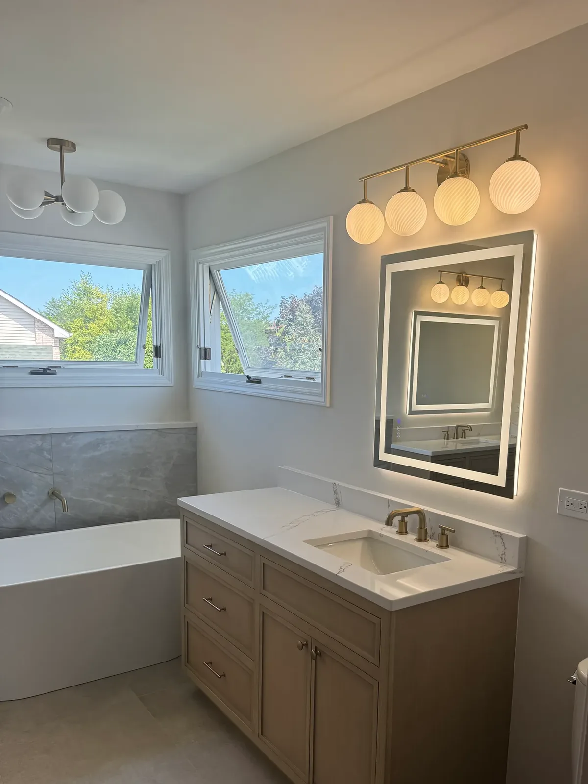Light-oak vanity with a white quartz top and a rectangular undermount basin under a backlit mirror and brass globe lighting, with the freestanding tub at left - Shorewood IL