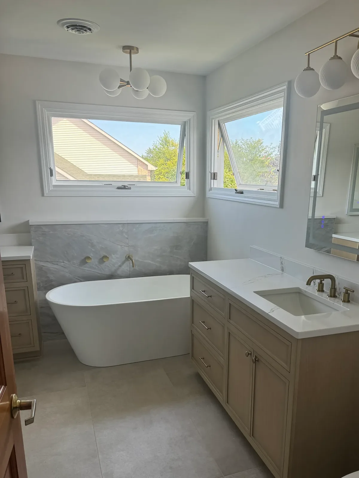 Primary bathroom in Shorewood, IL after renovation - freestanding tub against a grey marble-look porcelain wall beside a light-oak double vanity with a white quartz top, under two windows