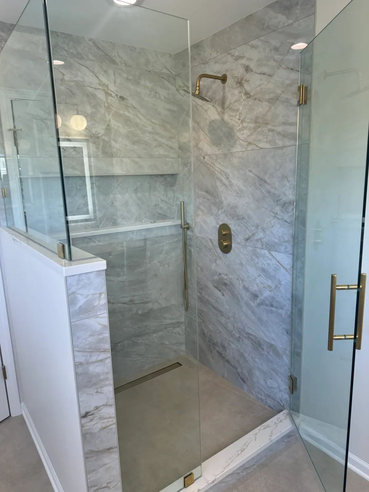 Looking into the primary shower in Shorewood, IL past a porcelain-clad knee wall - large-format grey marble-look porcelain, a full-width recessed niche, a brass rain head and a linear drain