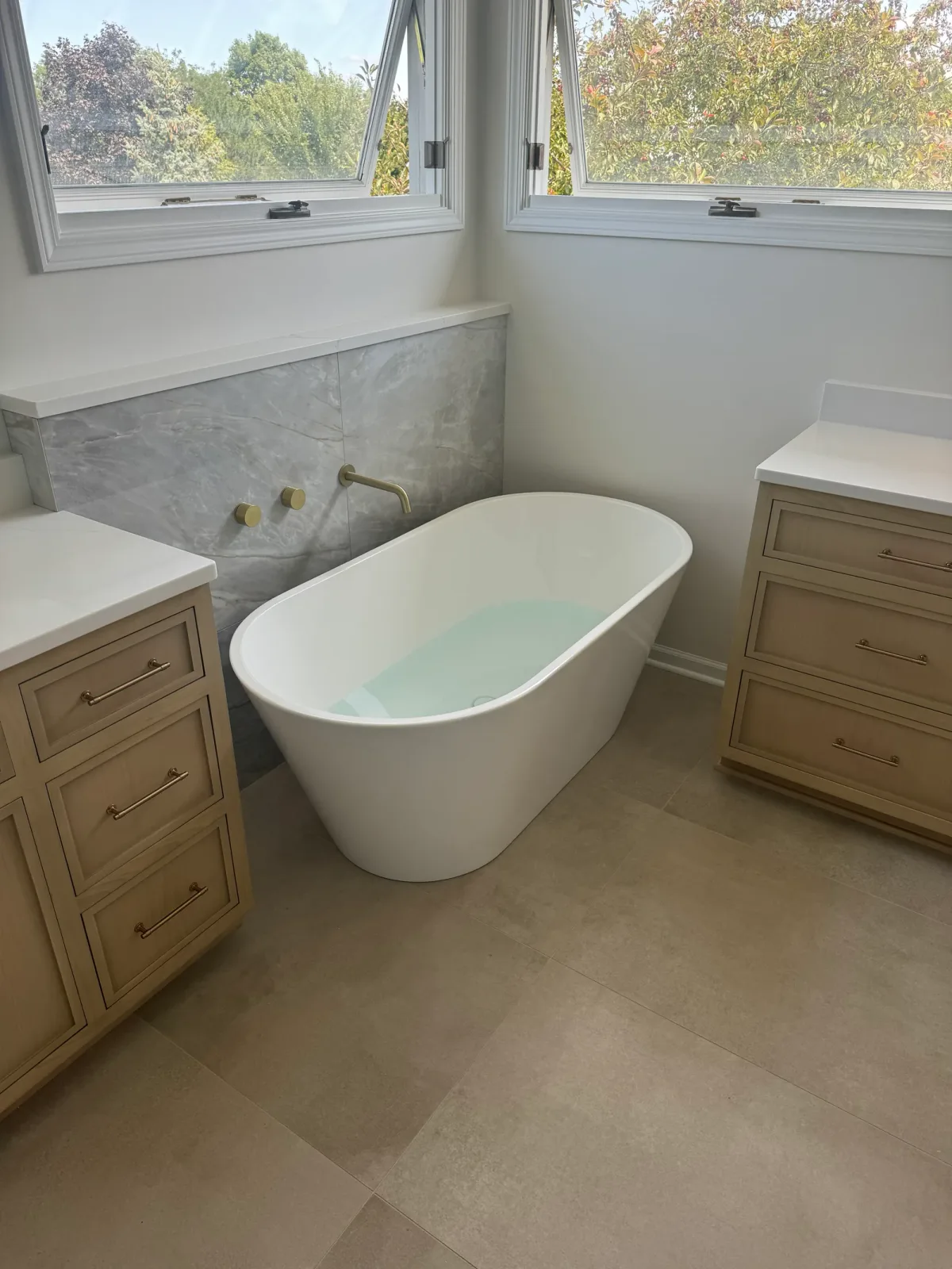 Freestanding soaking tub against a grey marble-look porcelain wall with a brass wall-mounted filler and two brass handles, flanked by light-oak cabinetry - Shorewood IL