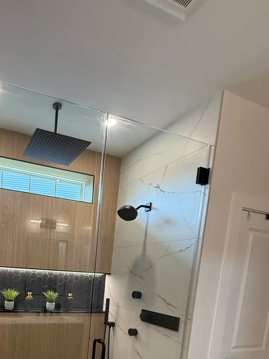 Bathroom remodel after photo, Shorewood IL - frameless glass shower on matte black hardware with a black ceiling rain head, a warm wood-look reeded back wall, marble-look large-format side walls, a full-width lit niche in charcoal hex and a charcoal hex mosaic floor