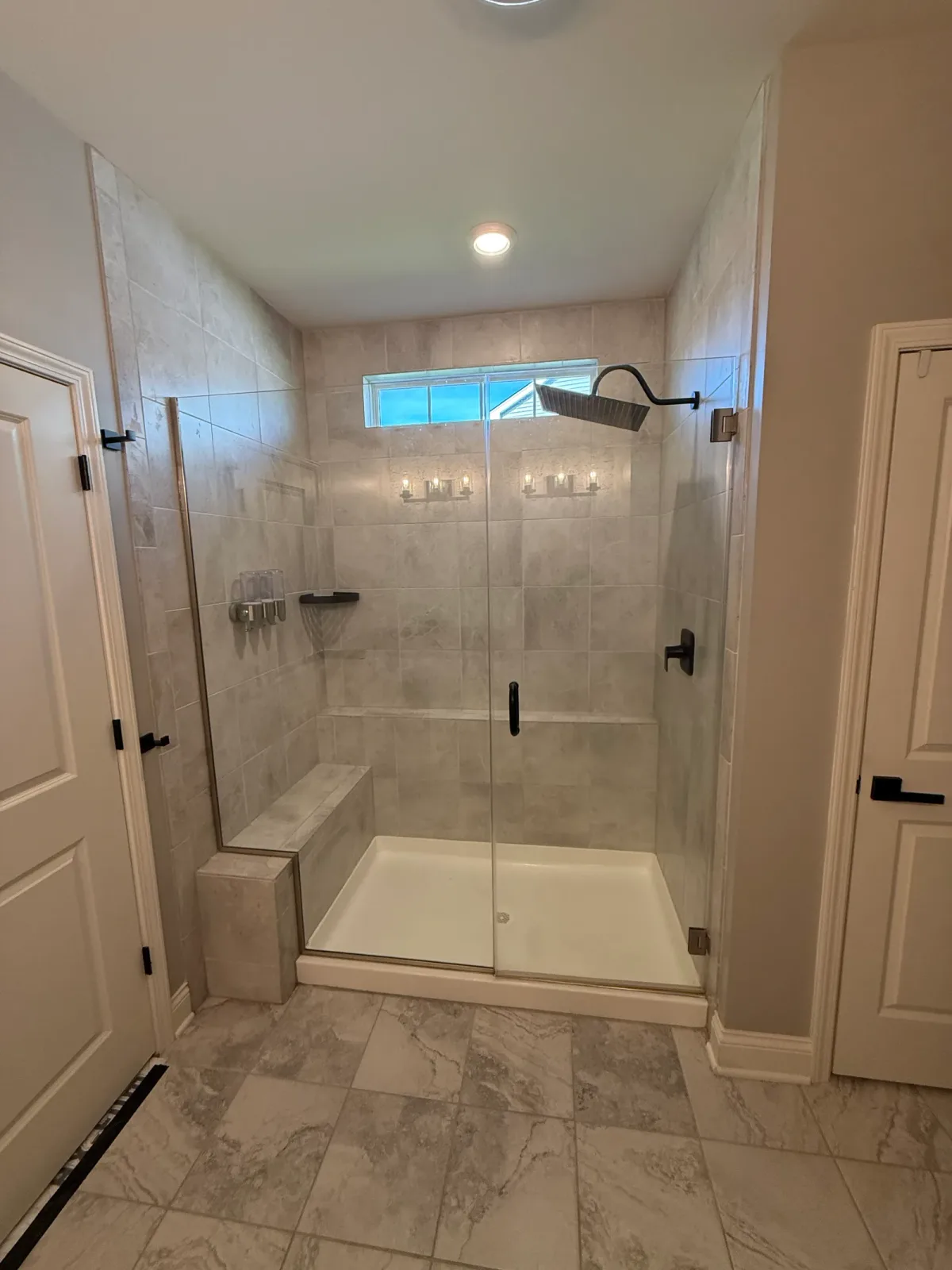 Bathroom remodel before photo, Shorewood IL - alcove shower in beige stone-look tile with a built-in tiled bench, a one-piece white pan with a center drain, glass panels in slim metal channels and a curved bronze arm with a square rain head under a transom window