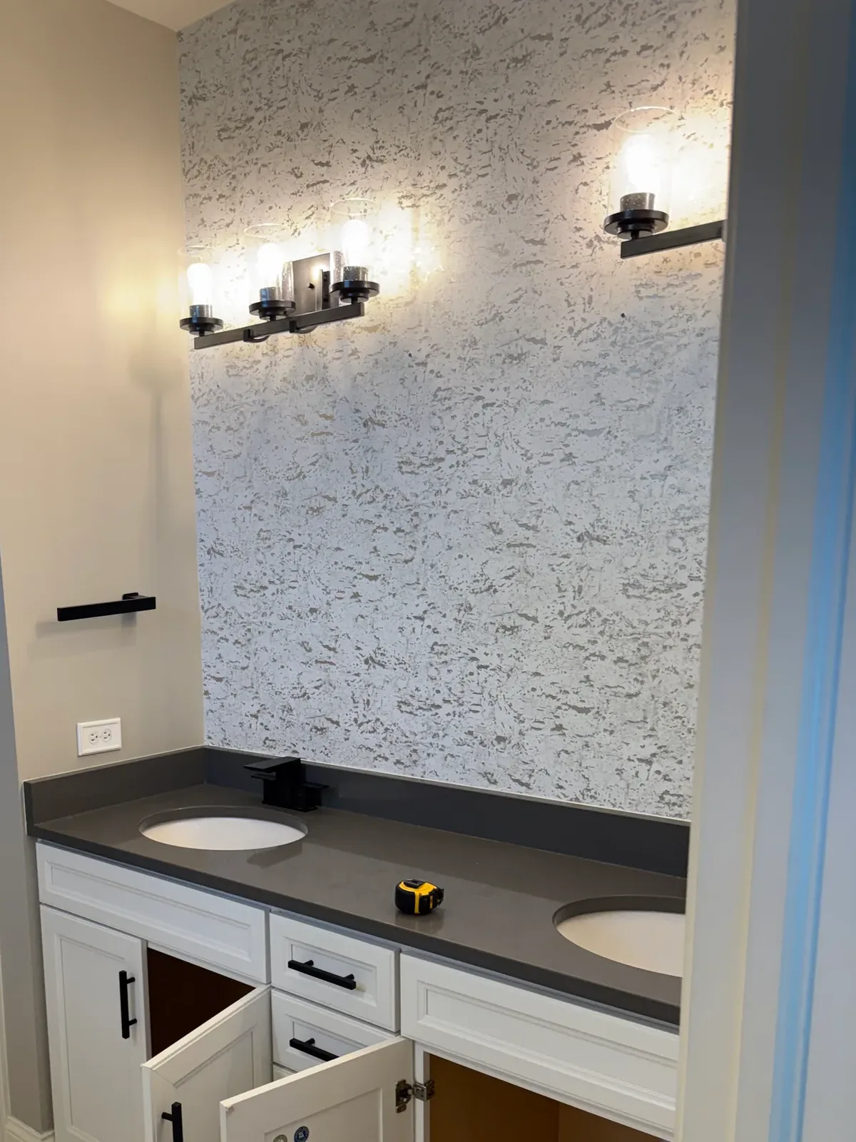 Bathroom remodel before photo, Shorewood IL - white raised-panel double vanity with a dark grey counter and two oval undermount bowls under a heavily textured white-and-silver-grey wall covering, lit by seeded-glass bar lighting in dark bronze