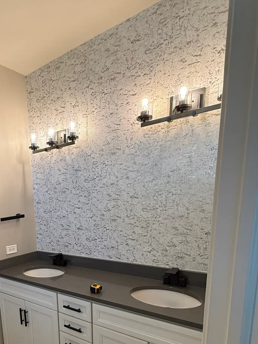 Bathroom before photo, Shorewood IL - a heavily textured white-and-silver-grey wall covering above a dark grey counter with two oval undermount bowls, lit by a pair of dark bronze three-light bars with seeded-glass shades