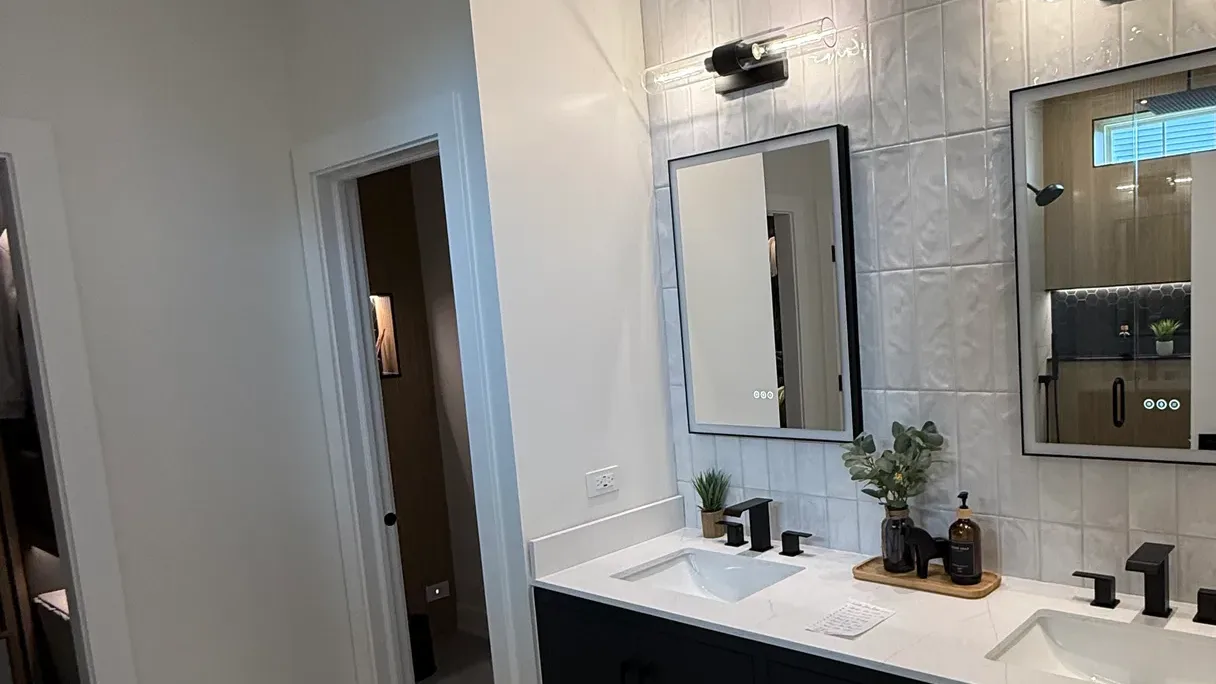 Bathroom remodel in Shorewood, IL with a black flat-panel double vanity, a white stone-look counter with rectangular basins, matte black tapware and two black-framed backlit mirrors on glossy white stacked tile by Luxe Development