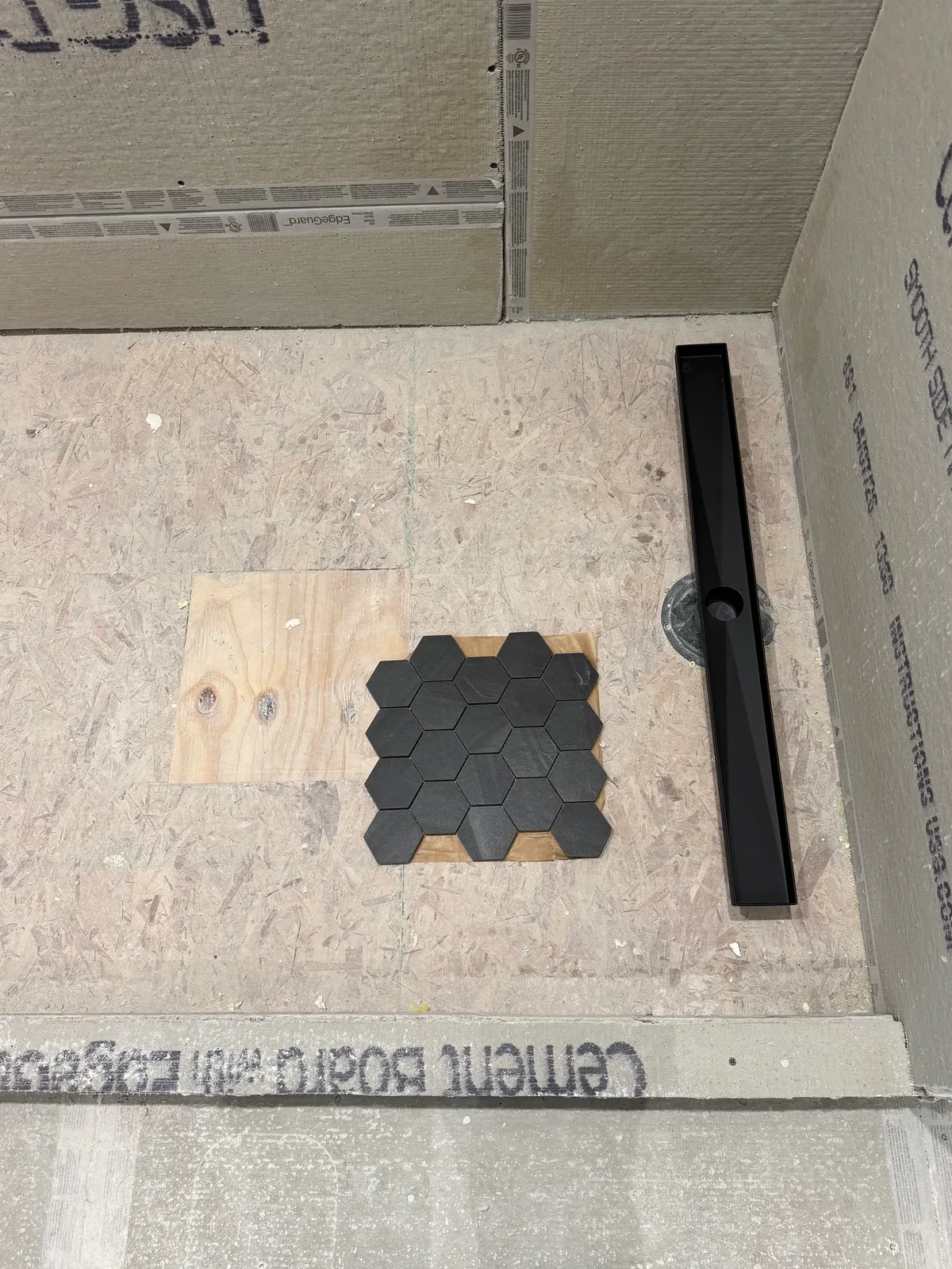 Shower rebuild in progress, Shorewood IL - cement board walls over a bare OSB subfloor, a matte black linear drain lying beside the exposed drain riser and a sheet of charcoal hex mosaic dry-laid on the subfloor