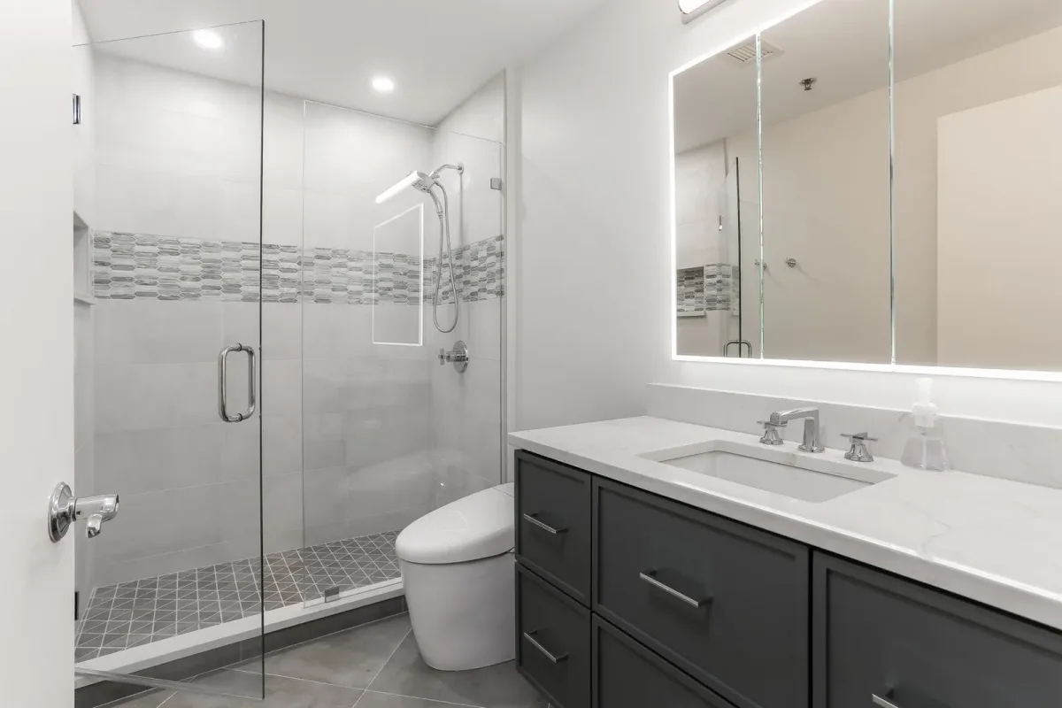 Second bathroom with a glass shower, mosaic accent band and a backlit mirror, condo renovation in Chicago, IL