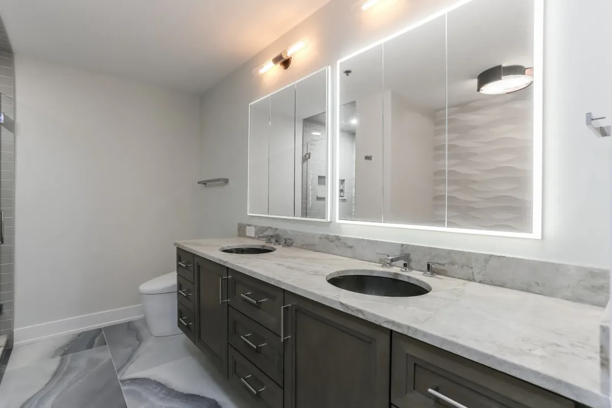 Dual undermount basins in a marble-veined slab top over stained cabinetry beneath backlit mirrors in a Chicago condo bathroom renovation