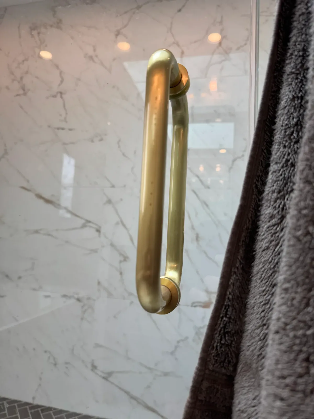 Close view of an unlacquered brass shower door pull against the marble wall, with a folded towel behind - marble primary bath remodel, Mokena IL
