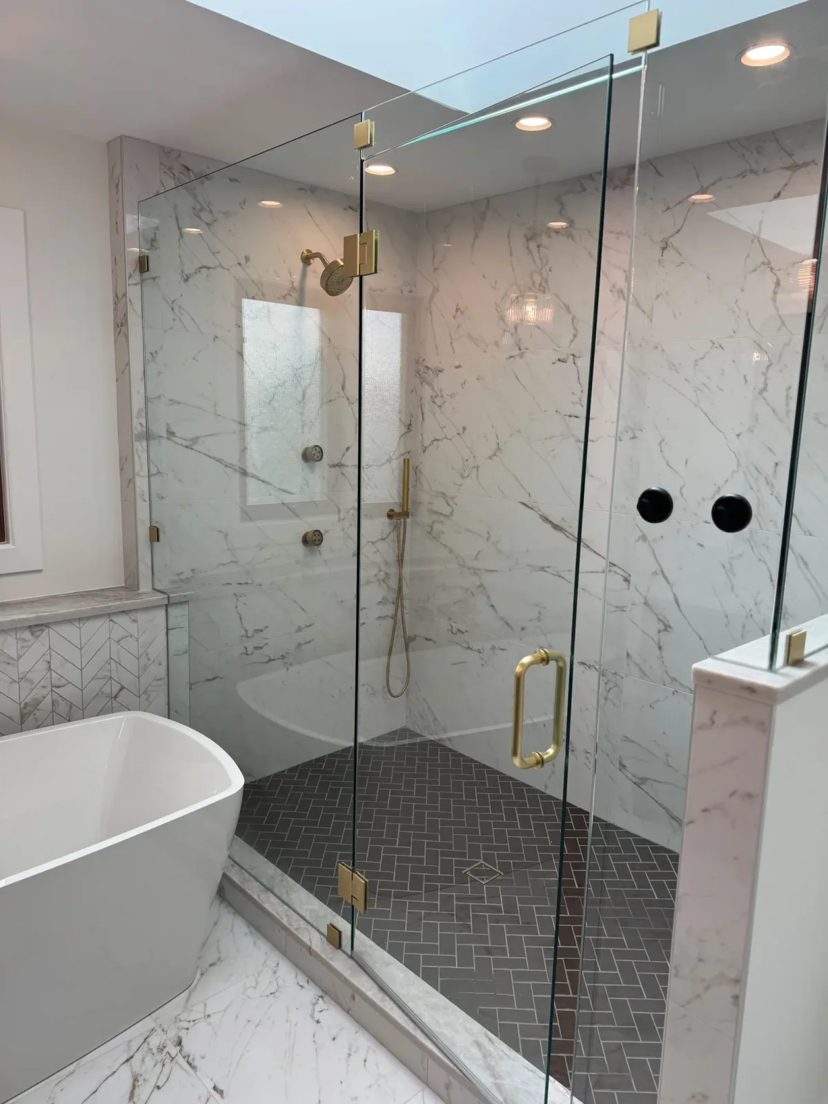 Frameless glass shower enclosure seen past the freestanding tub, with the marble wall and dark herringbone floor beyond - marble primary bath remodel, Mokena IL