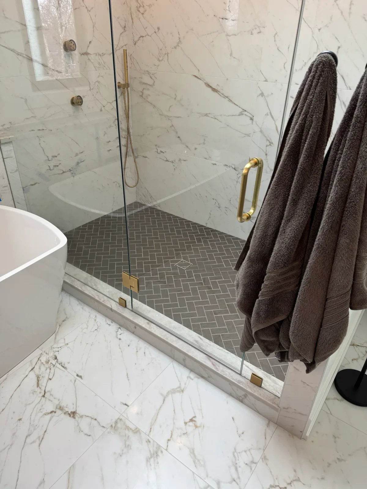 Frameless glass shower with a dark herringbone tile floor, a brass door pull and folded towels on a brass rail - marble primary bath remodel, Mokena IL