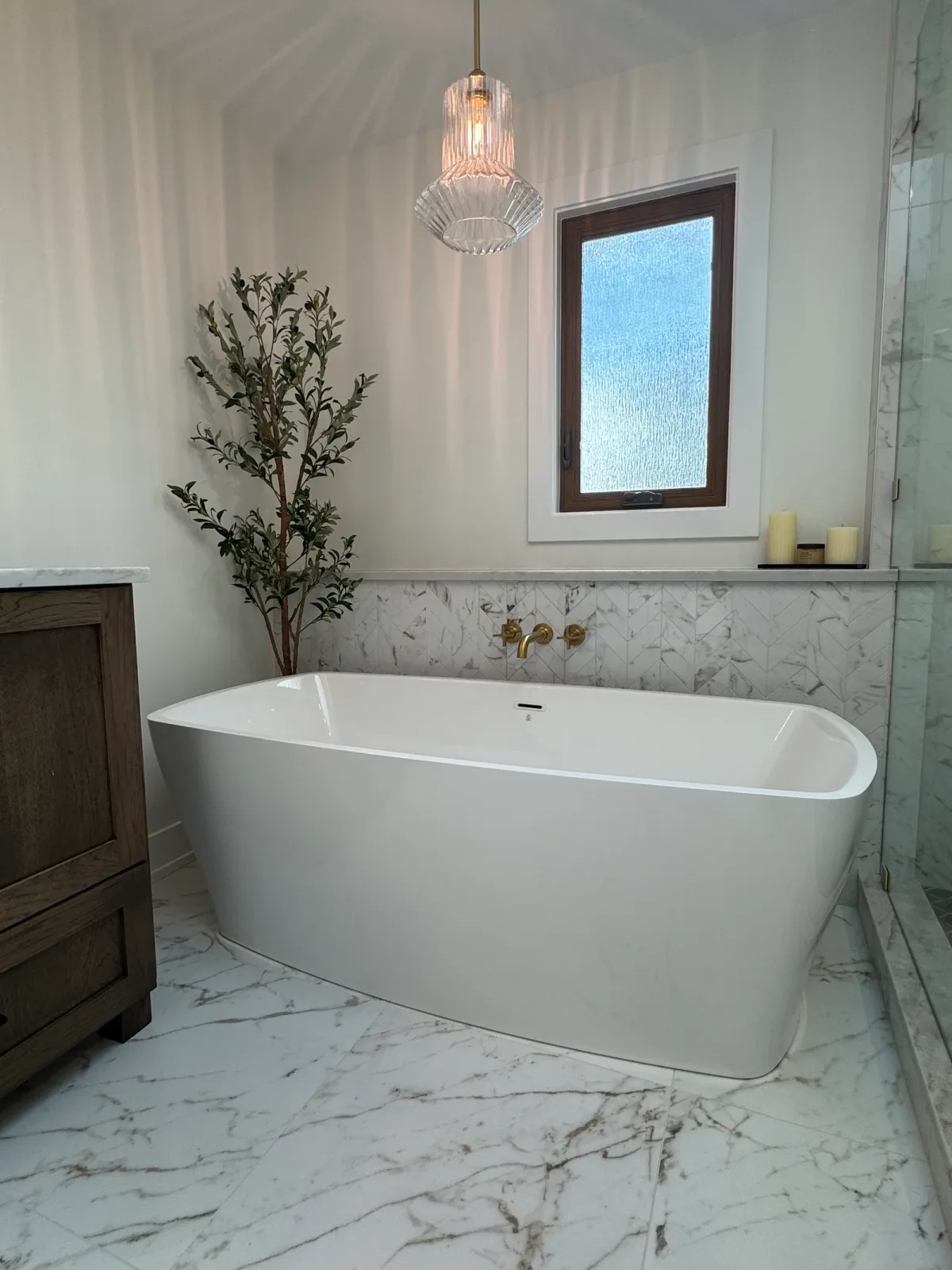 Freestanding soaking tub against a marble-clad wall with a floor-mounted brass filler and an olive tree beside it - marble primary bath remodel, Mokena IL