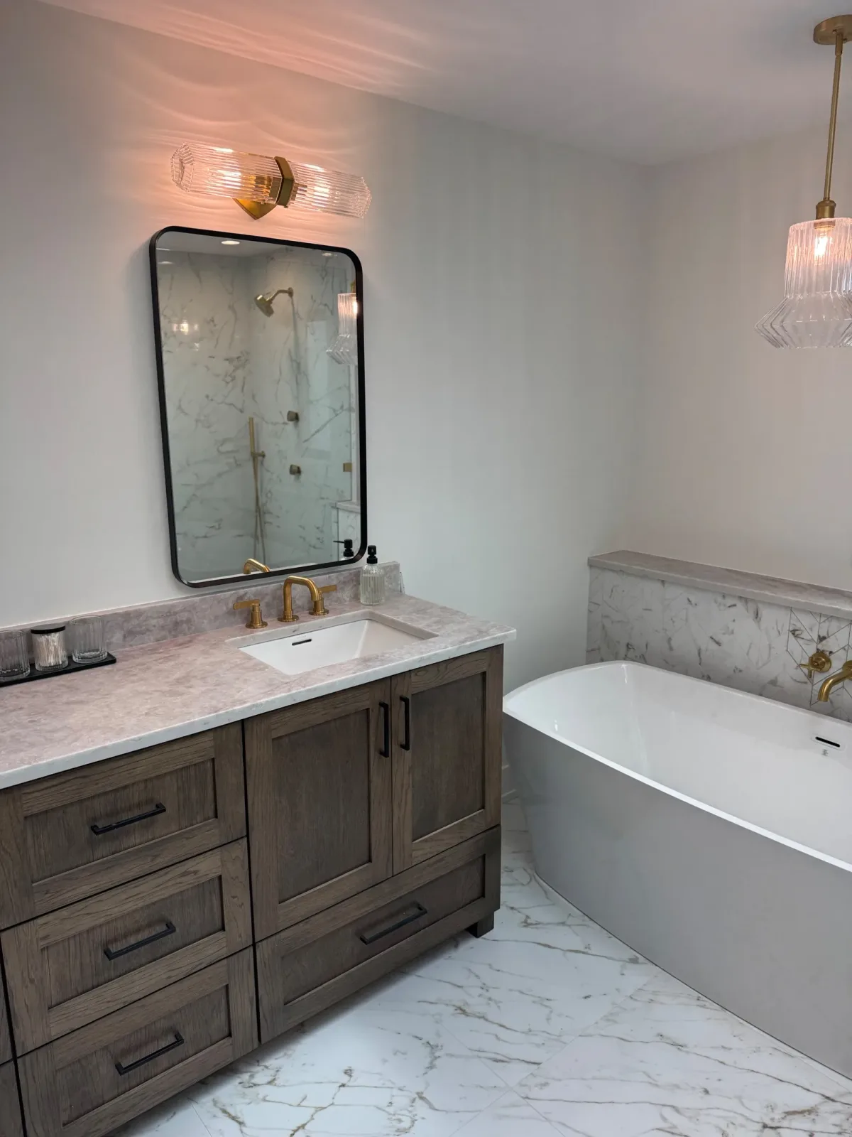 Stained oak double vanity beside the freestanding tub, with brass sconces above the mirrors and marble underfoot - marble primary bath remodel, Mokena IL