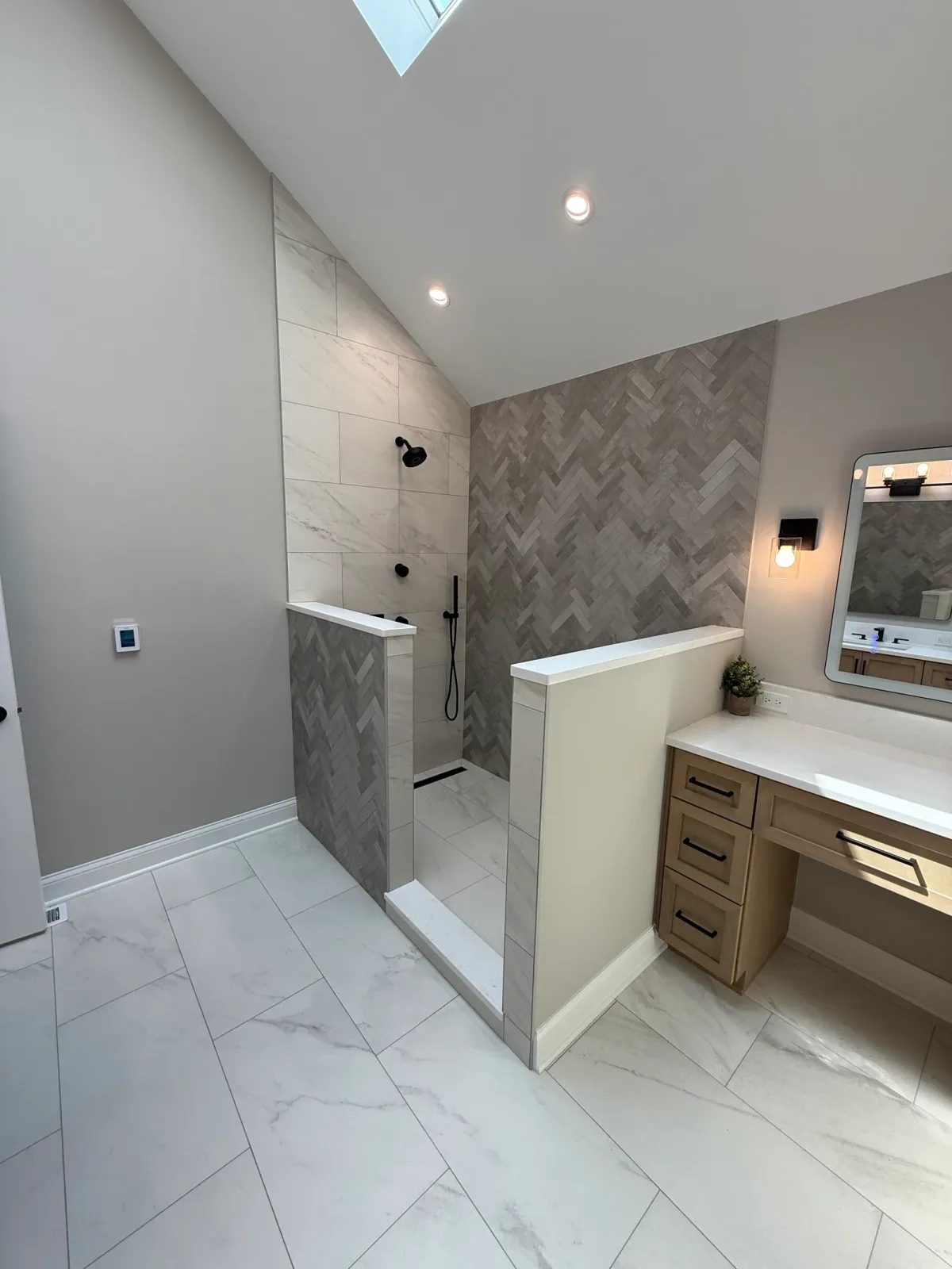 Wide view of the open walk-in shower, its knee walls, the herringbone accent wall and the tiled floor, with the seated makeup station at the right edge - primary bath remodel, Mokena IL