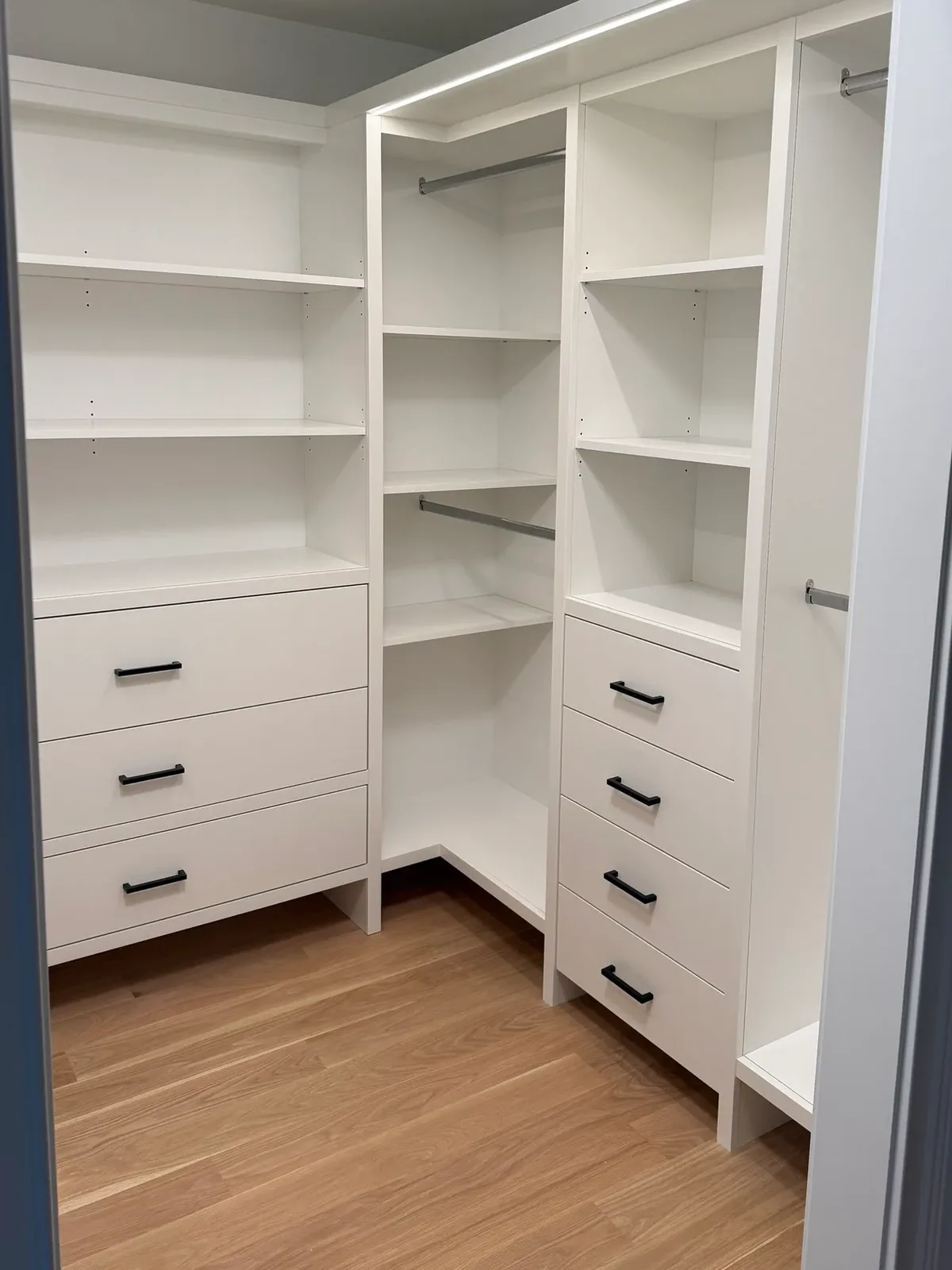 White painted walk-in closet built-ins with adjustable shelving, banks of drawers on black bar pulls, chrome hanging rods and an LED-lit top shelf, over a white oak floor - whole-home renovation, Mokena IL