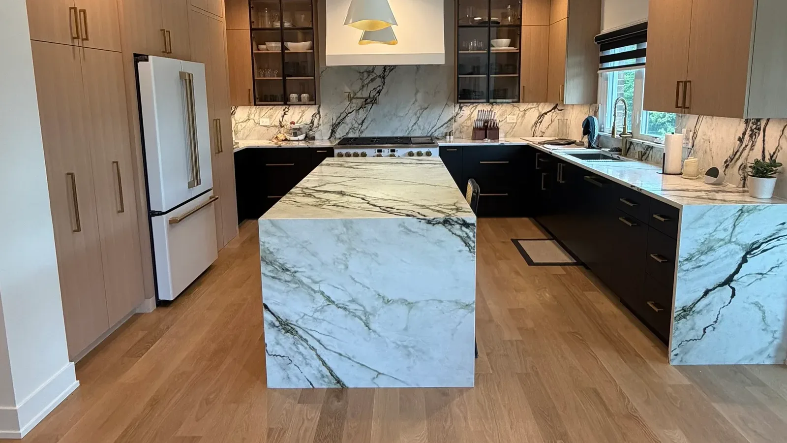 Whole-home renovation in Mokena, IL by Luxe Development - kitchen with a heavily veined stone island whose top turns the corner and runs to a white oak floor, white oak and matte black cabinetry, a full-height stone backsplash behind the range and brass hardware