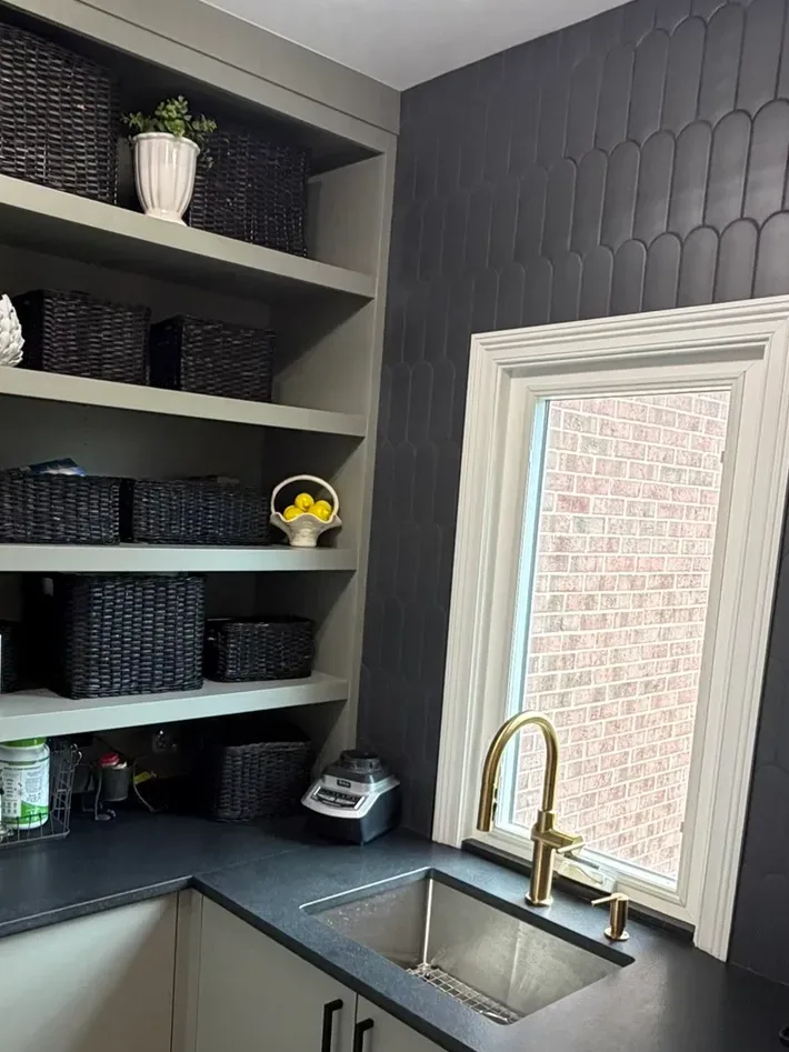 Prep pantry with a charcoal fan-shaped tile wall, a brass gooseneck tap over a stainless undermount sink, a black stone counter and sage-painted open shelving stocked with woven baskets - whole-home renovation, Mokena IL