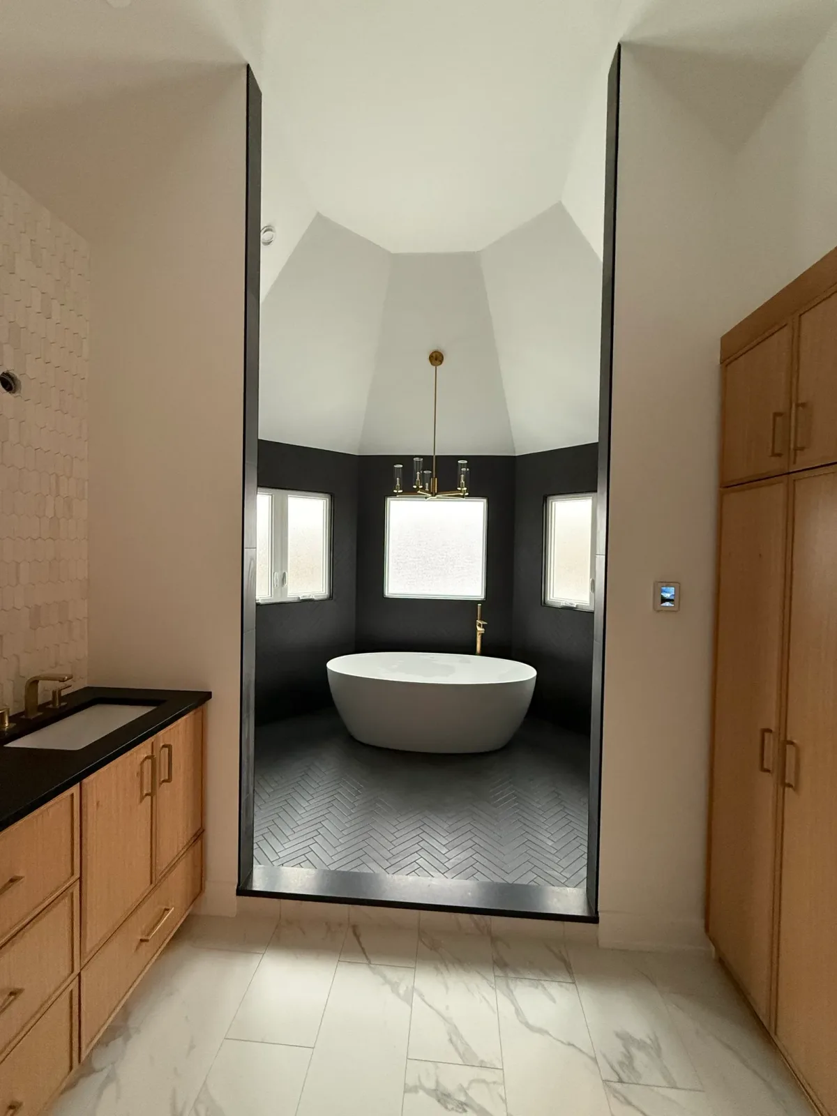 Primary bathroom in Mokena, IL with a white oak vanity and black stone counter at left, a tall white oak linen tower at right, a marble-look large-format tile floor, and a dark-tiled opening into a matte black herringbone tub alcove beneath a white vaulted ceiling
