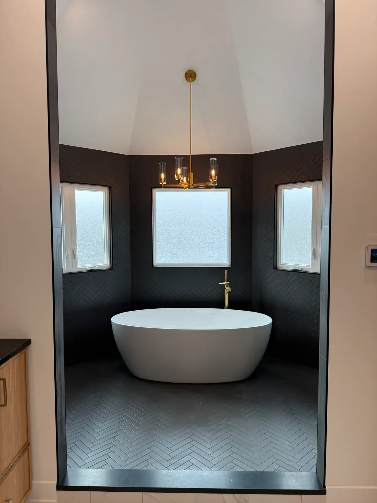 Freestanding oval soaking tub in an alcove finished in matte black herringbone tile on the walls and floor beneath a white vaulted ceiling, with a brass floor-mounted filler, a brass chandelier and three obscure-glass windows - whole-home renovation, Mokena IL