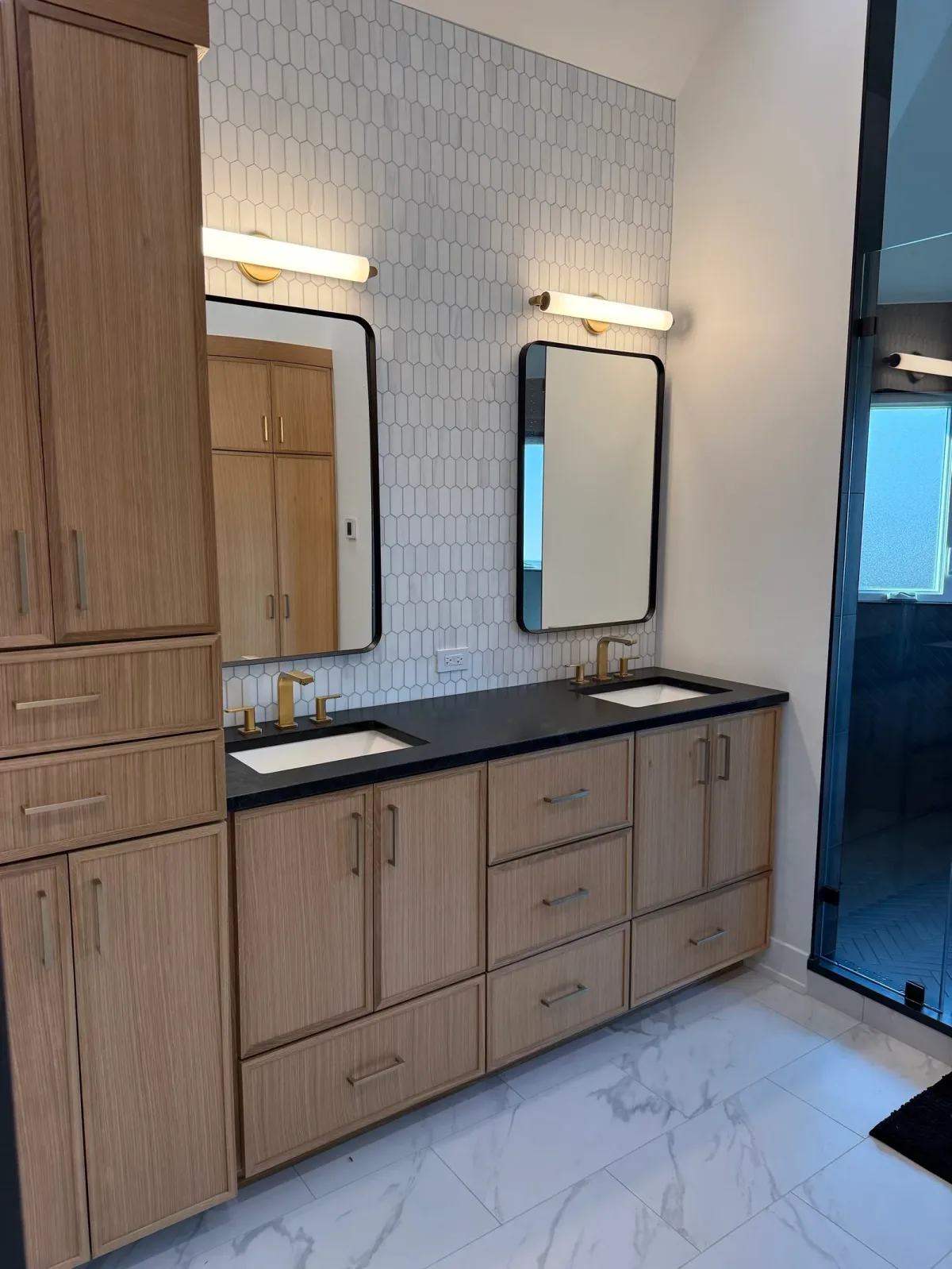 White oak double vanity with a black stone counter, white undermount basins and brass widespread faucets against a warm-white picket tile wall, with black-framed mirrors and brass-mounted sconces above - whole-home renovation, Mokena IL